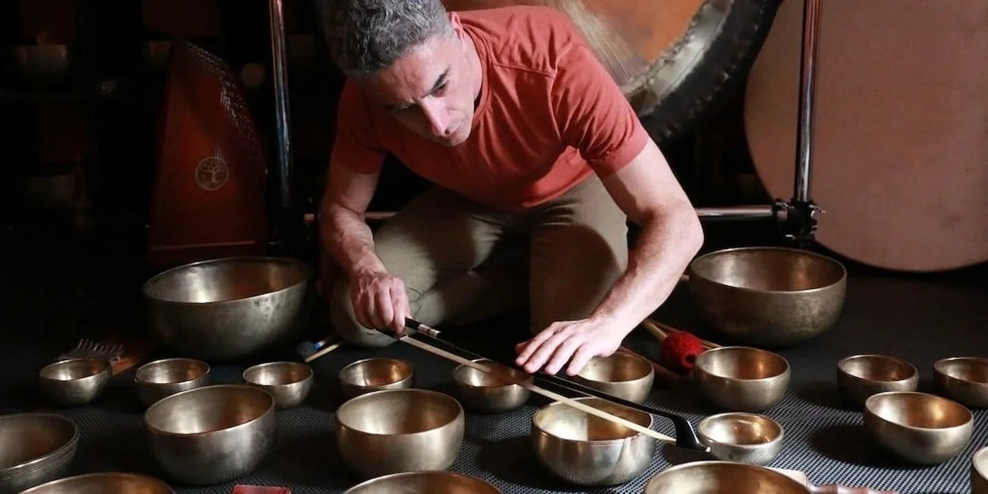 A man in a red shirt and beige pants is sitting on the floor surrounded by singing bowls, holding a mallet and playing one of the bowls.