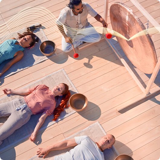 People lying down on yoga mats during a sound healing session with a gong and singing bowls, led by a person playing a mallet in a bright room with wooden flooring.
