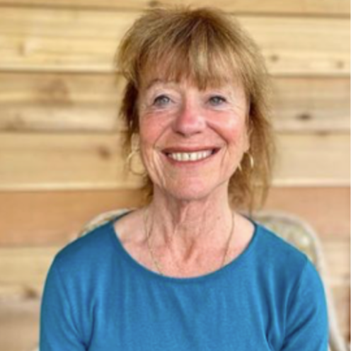 A smiling elderly woman with short, light brown hair wearing a blue top, sitting against a wooden wall background.