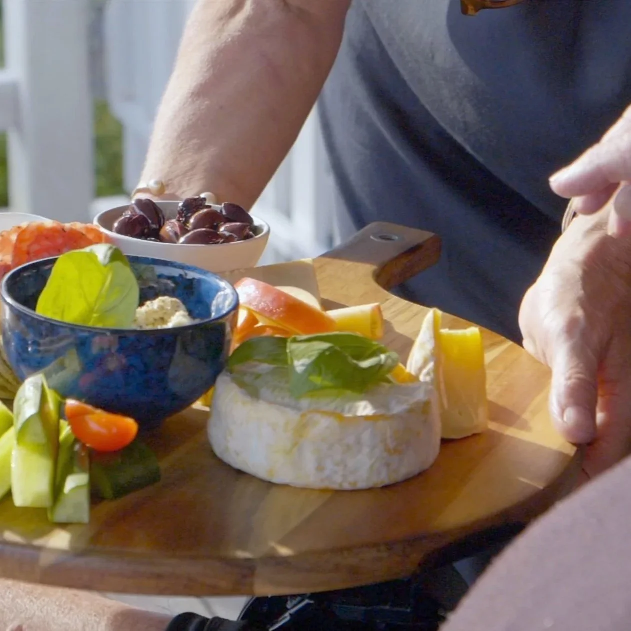 Person slicing brie cheese on a wooden cutting board with assorted cheeses, grapes, and vegetables nearby.
