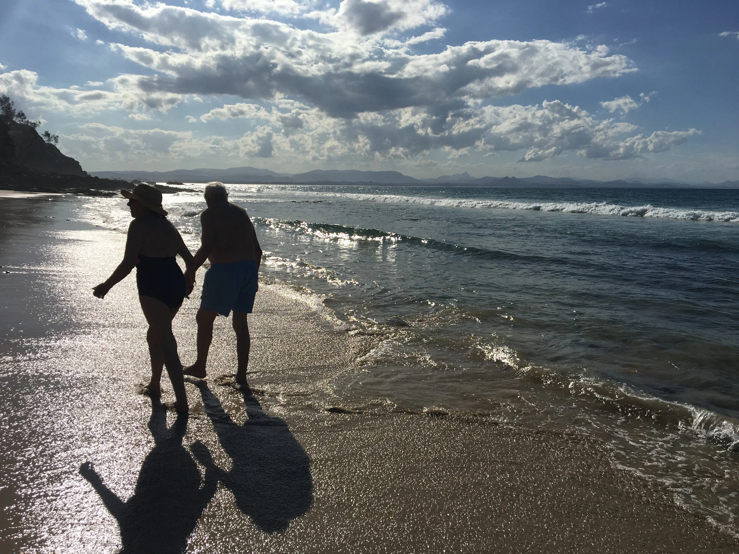 An elderly couple walking hand in hand along the beach at sunset or late afternoon, with the ocean waves gently rolling in and a partly cloudy sky.