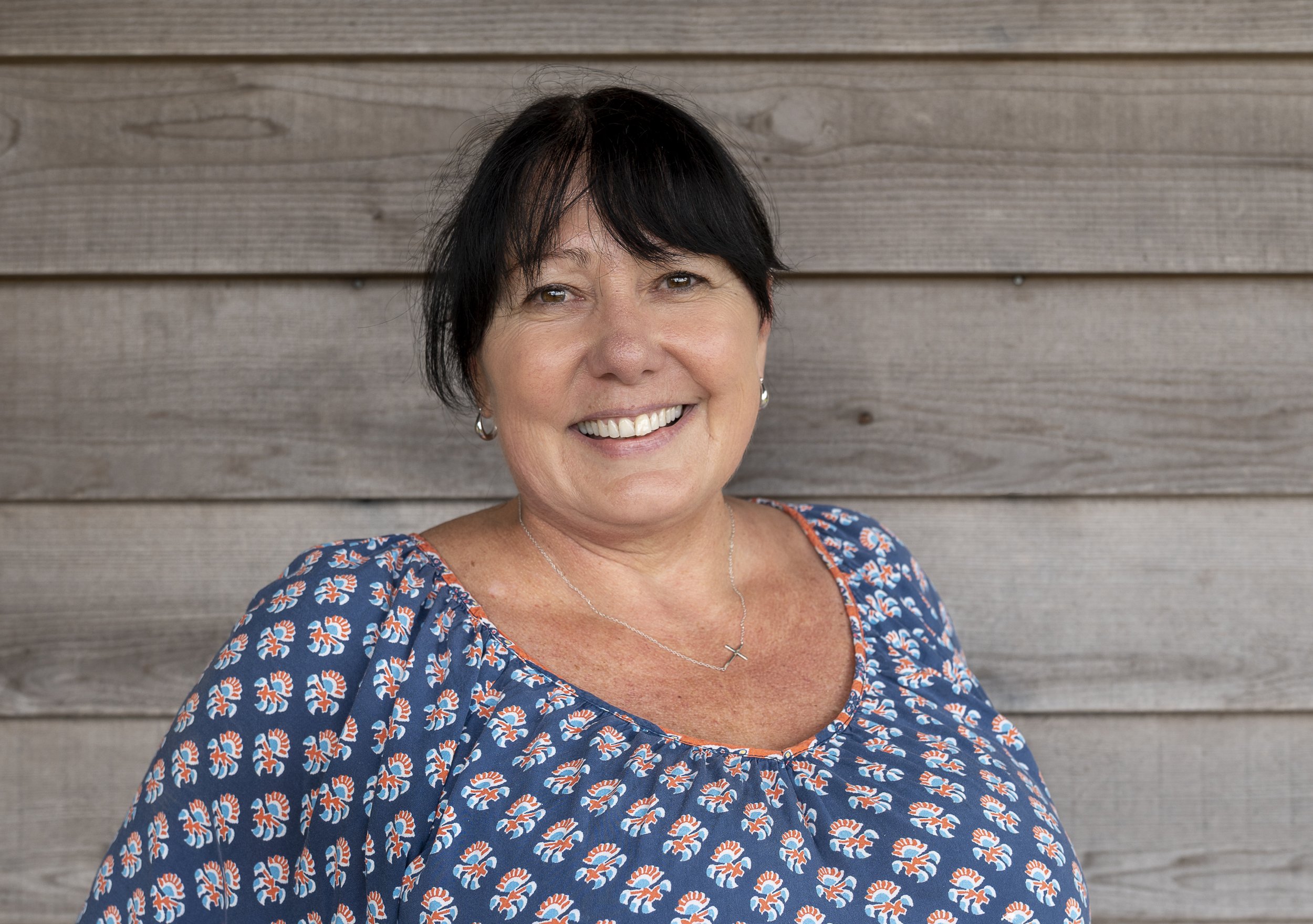 A woman with short dark hair, wearing a pink scarf and a patterned sweater, smiling in front of a wooden wall.