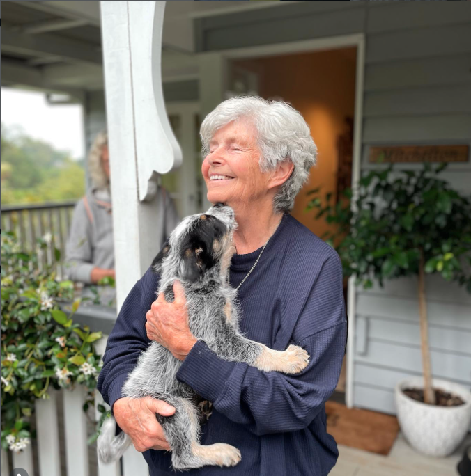 An elderly woman with white hair smiling and hugging a black and white puppy on a porch with greenery and a person in the background.