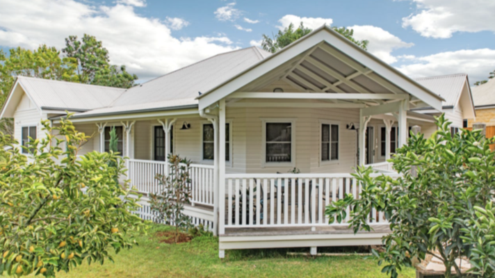 White house with a porch and a metal roof, surrounded by green trees and shrubs under a partly cloudy sky.