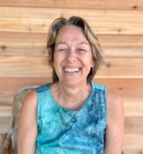 A woman with short gray hair smiling, wearing a sleeveless blue patterned top, sitting in front of a wooden wall.