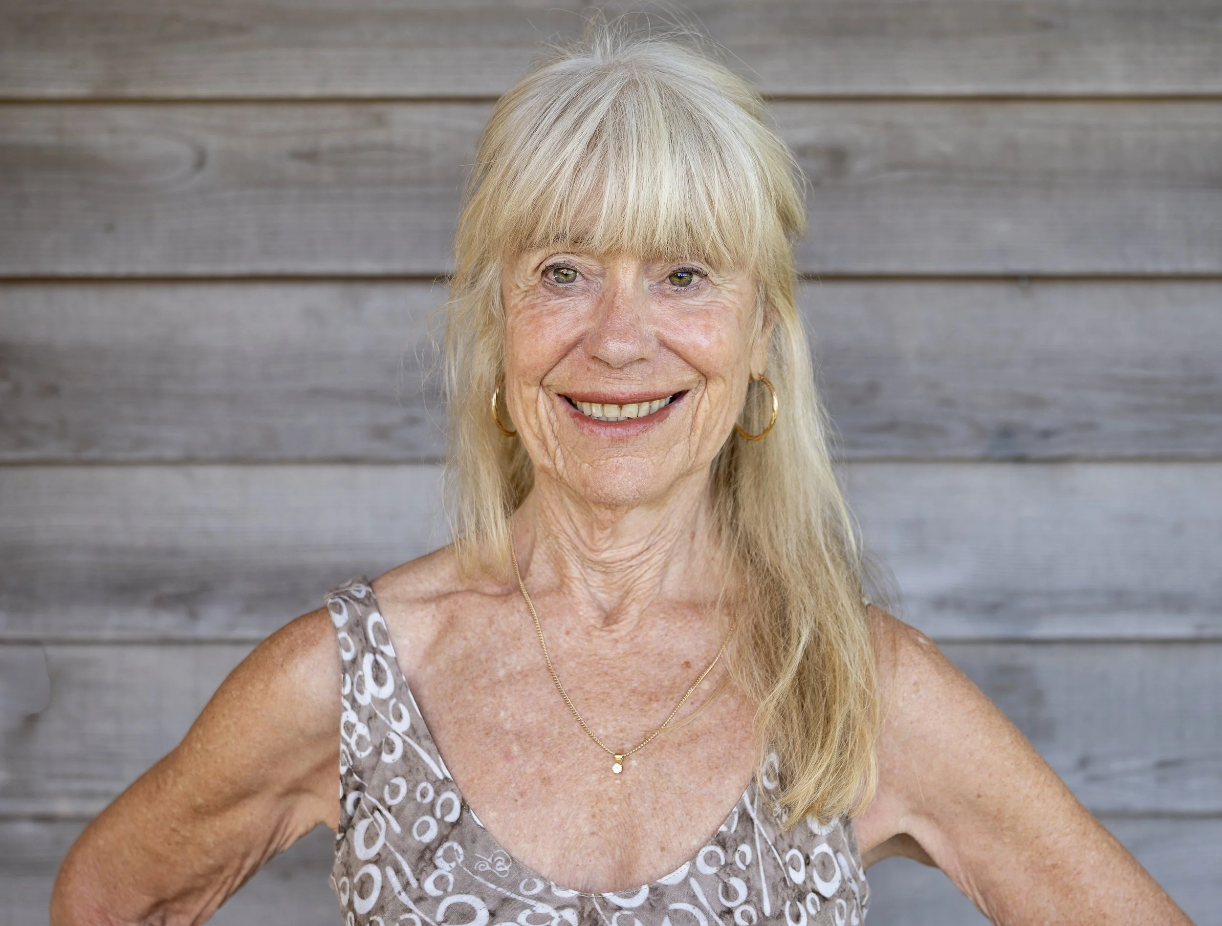 A smiling elderly woman with short, light brown hair wearing a blue top, sitting against a wooden wall background.