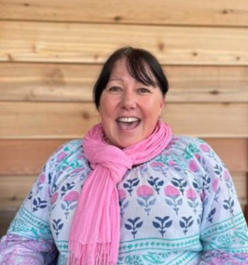 A woman with short dark hair, wearing a pink scarf and a patterned sweater, smiling in front of a wooden wall.