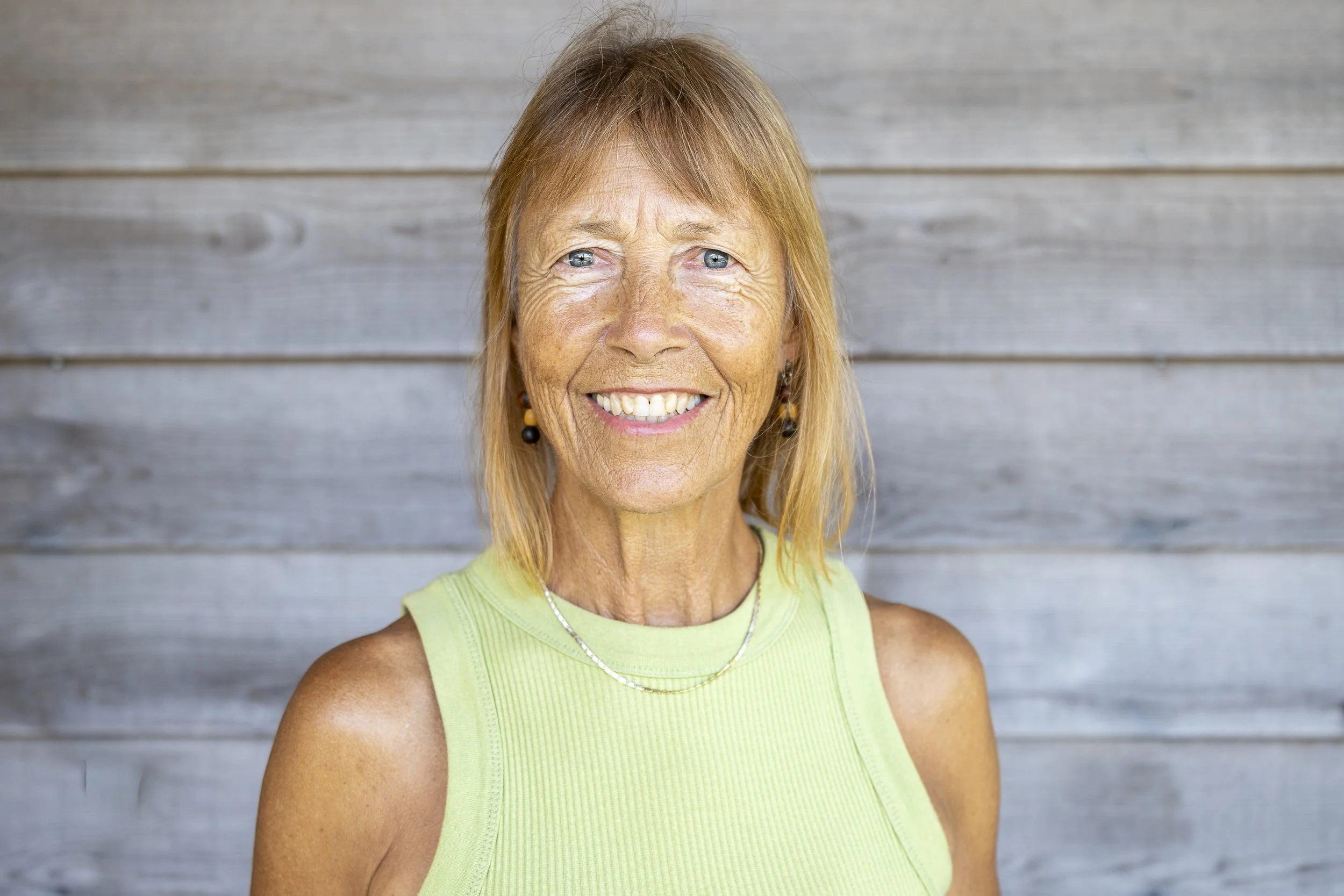 A smiling woman with short blonde hair sitting on a chair with a wooden wall background.