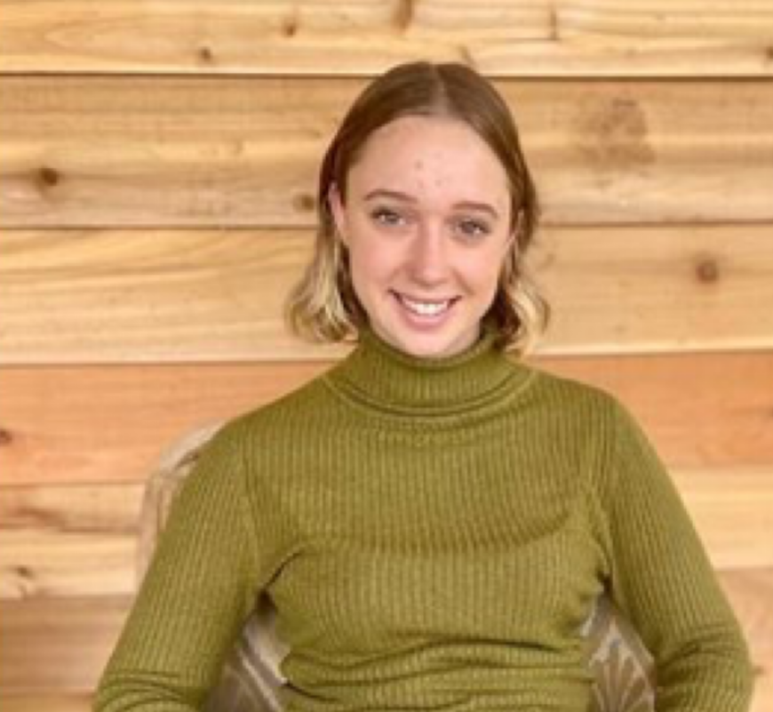 A woman with shoulder-length blonde hair, wearing an olive green turtleneck sweater, sitting against a background of wooden paneling.
