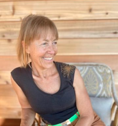 A smiling woman with short blonde hair sitting on a chair with a wooden wall background.