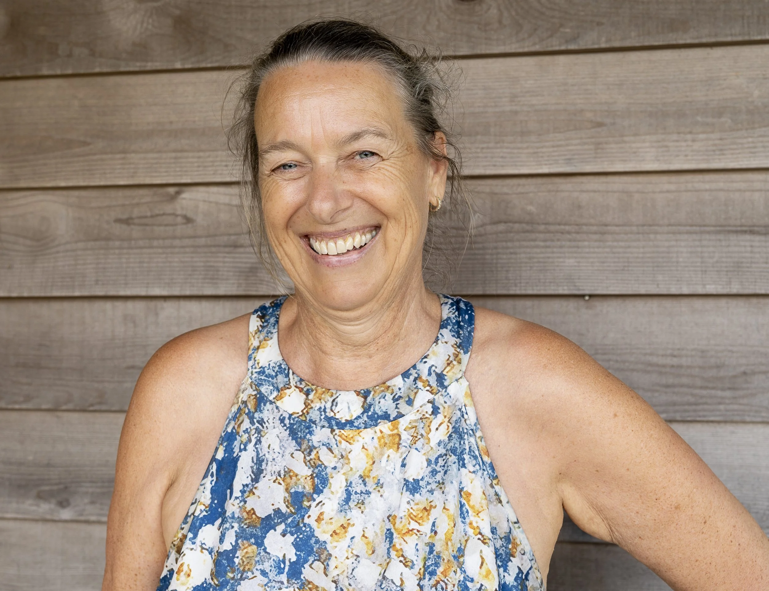 A woman with short gray hair smiling, wearing a sleeveless blue patterned top, sitting in front of a wooden wall.