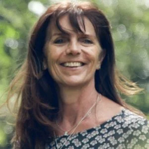A smiling woman with dark brown hair standing outdoors with green trees in the background.