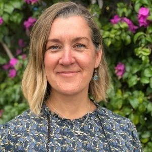 A woman with shoulder-length blonde hair and earrings standing outdoors in front of green foliage and pink flowers.