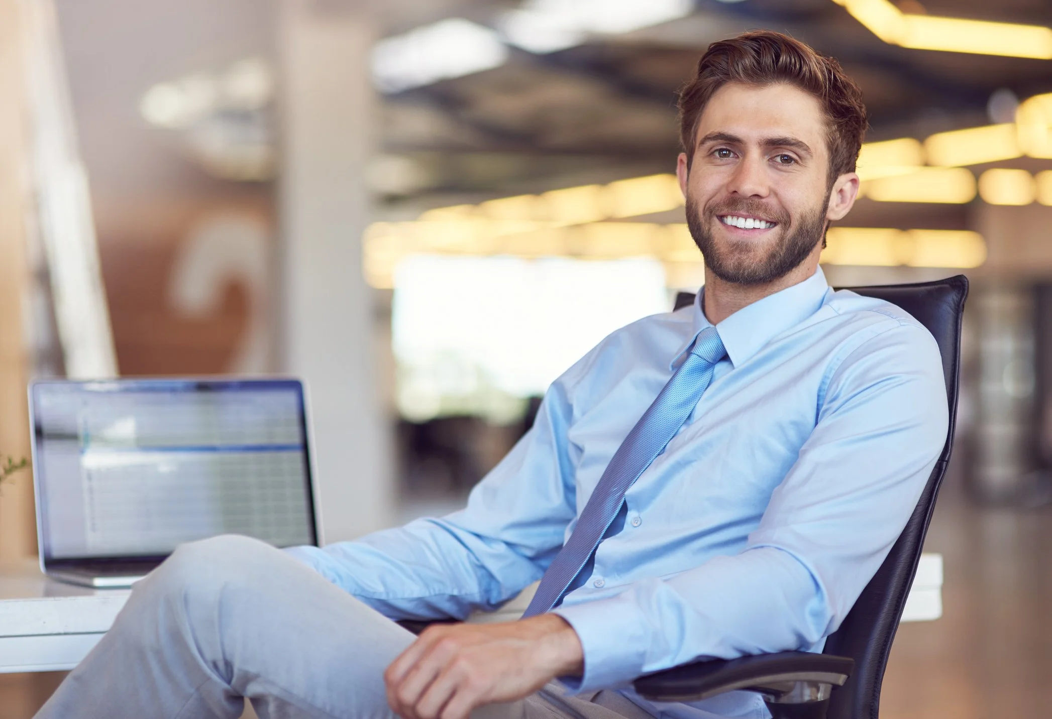 A smiling man sitting at a desk in an office, wearing a light blue dress shirt and a blue tie, with a laptop in front of him and blurred office background.