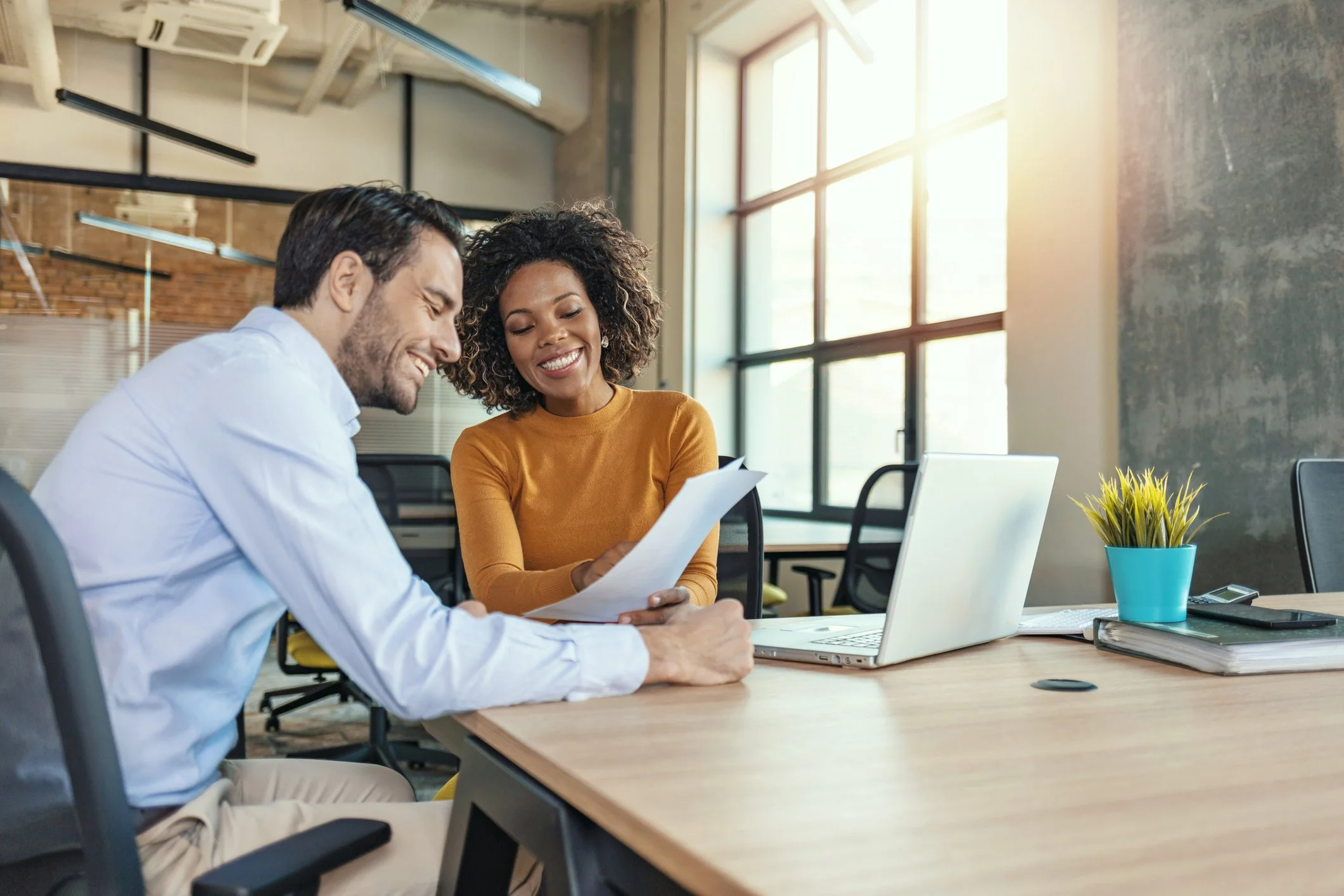 Two smiling colleagues, a man and a woman, are sitting at a desk in a modern office looking at a document together. The office has large windows and a laptop, a plant, and various office supplies on the desk.