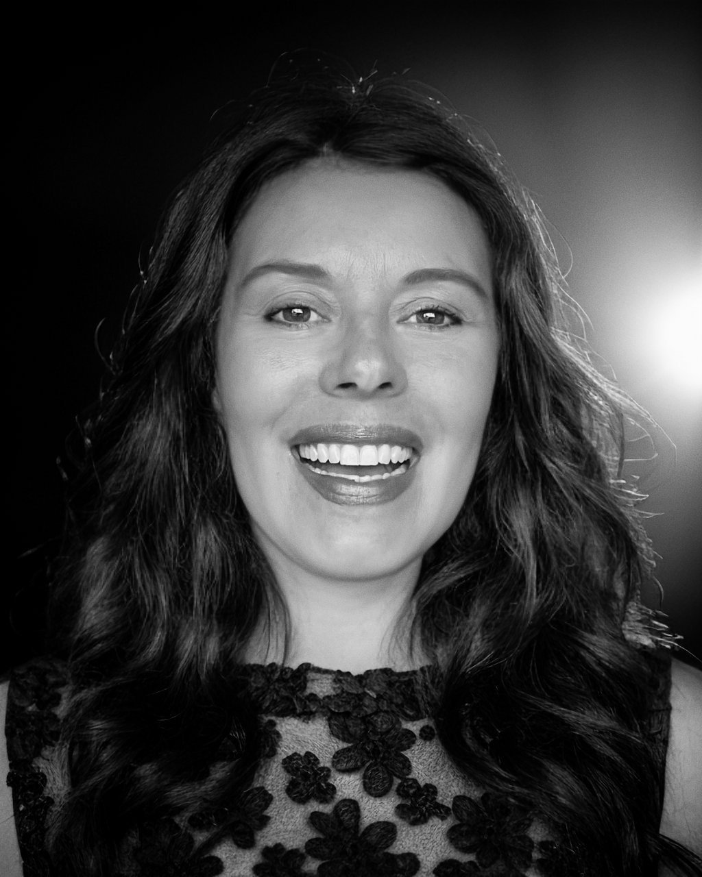 A smiling woman with long wavy hair, wearing a lace top, in black and white.