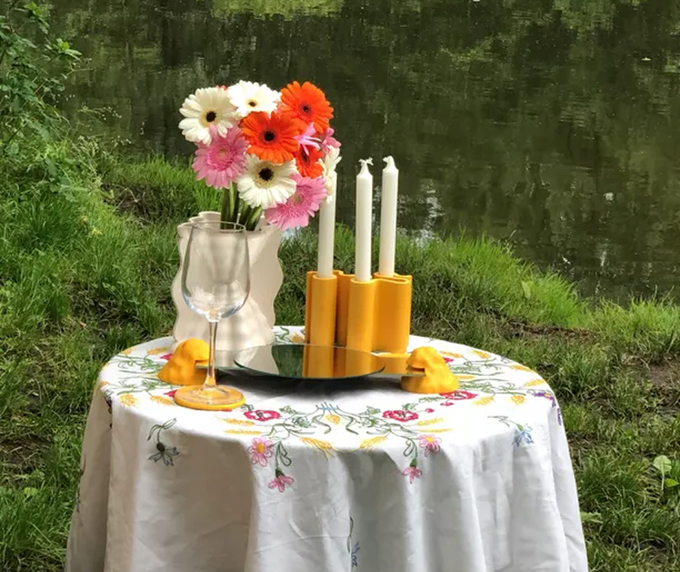 Table setup outdoors with a floral tablecloth, a glass vase with colorful flowers, a wine glass, three white candles in yellow holders, a reflective round tray, and two orange decorative items, near a body of water.