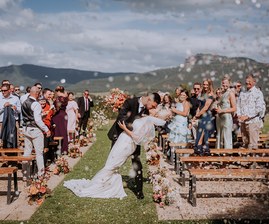 Bride and groom kissing during outdoor wedding ceremony with guests watching in a scenic mountain setting.