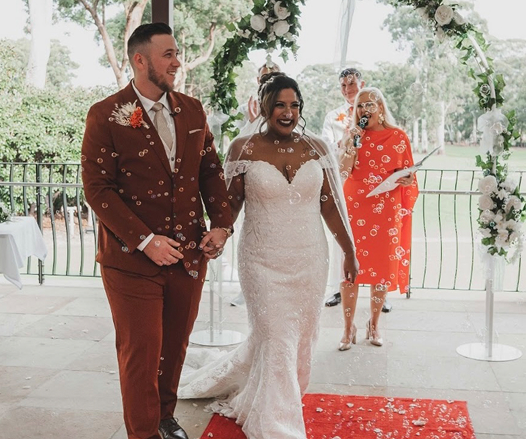 Bride and groom holding hands, smiling, standing on a red carpet, with an officiant in the background at an outdoor wedding ceremony.