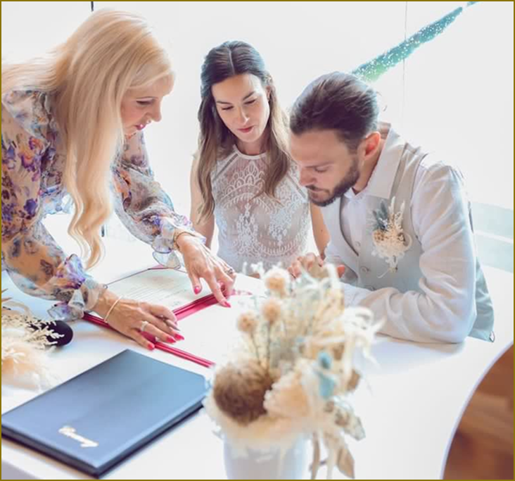 Three people, two women and one man, look at a book or photo album on a table. The table has a blue notebook and decorative dried flowers in a white vase. The scene suggests they are planning or reviewing something together.