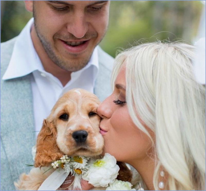 A woman with platinum blonde hair kisses a golden retriever puppy on the nose, while a man smiles in the background. The puppy has a white flower collar.