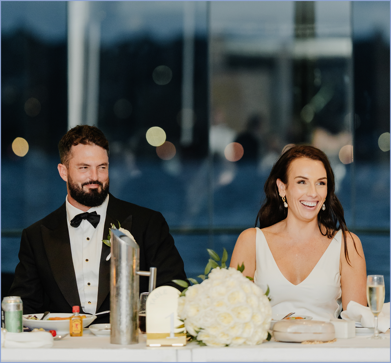 A bride and groom sitting at a wedding reception table, smiling and looking to the right. The bride has long dark hair and is wearing a white wedding dress, earrings, and makeup. The groom has a beard, short hair, and is wearing a black tuxedo with a bow tie. The table has a bouquet of white roses, champagne, and other drinks.
