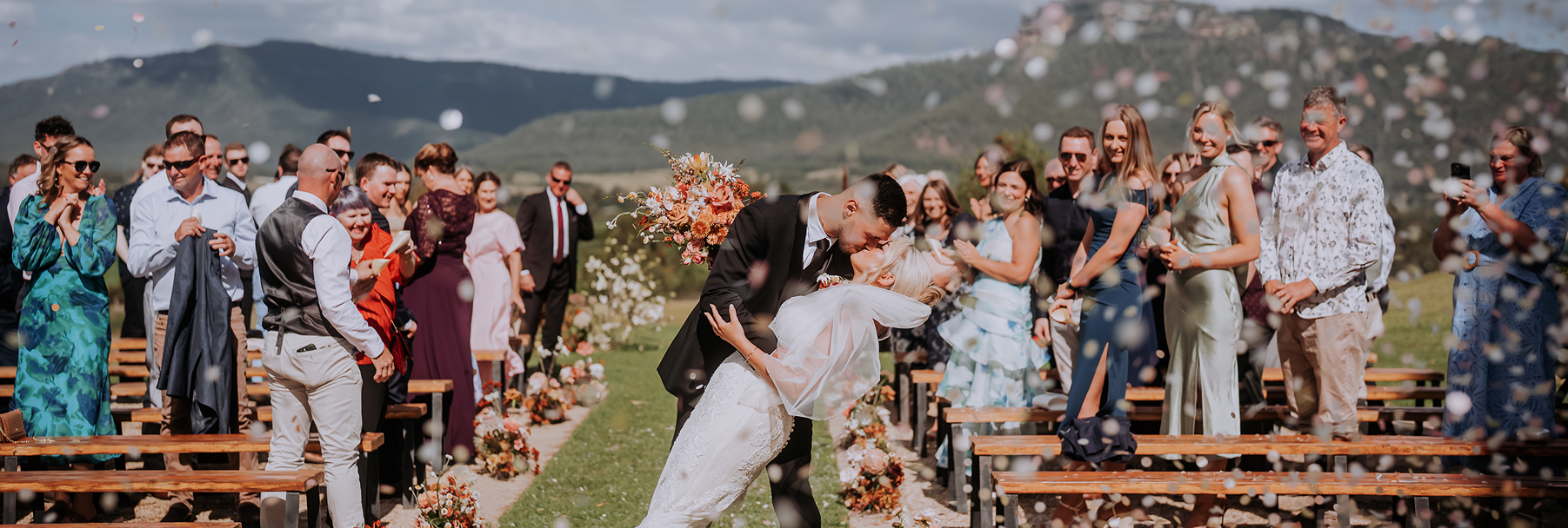 Bride and groom kissing at outdoor wedding ceremony with guests celebrating, mountain landscape in background.