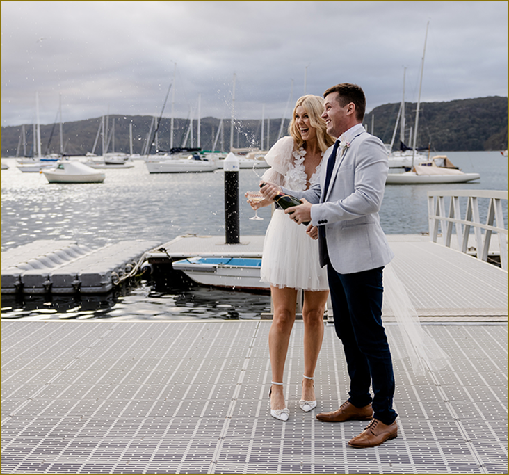 A couple celebrating on a dock at a marina with sailboats in the background, with the man opening a bottle of champagne.