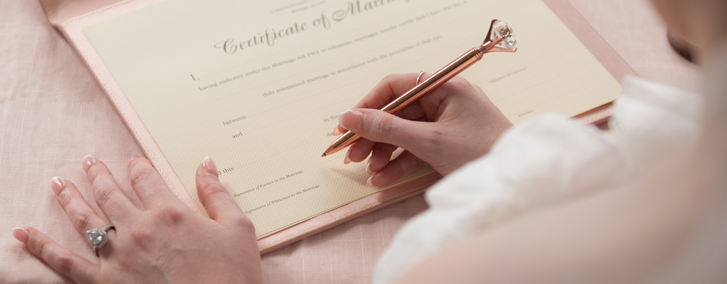 A person signing a wedding certificate with a decorative pen. The certificate is on a pinkish folder, placed on a pink fabric surface.