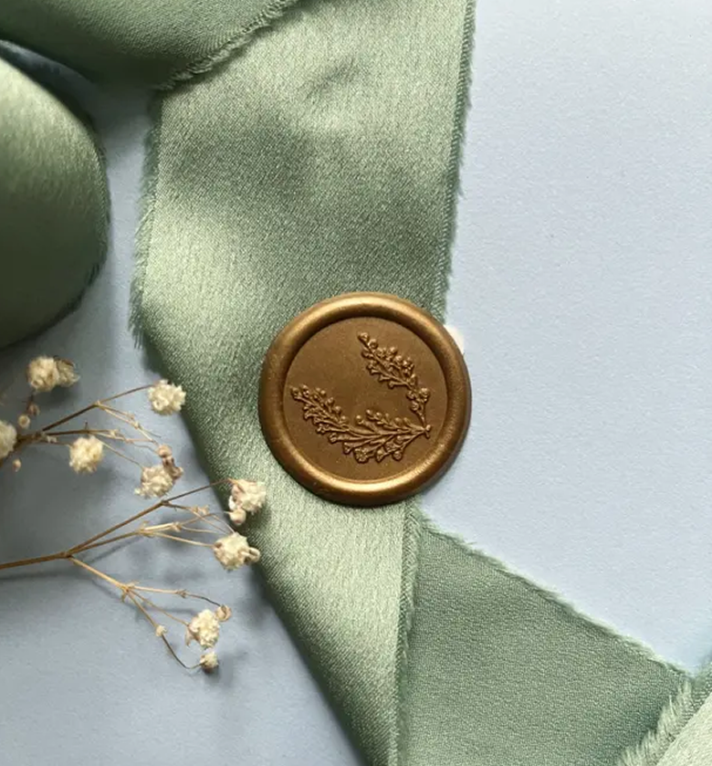 Close-up of a light green satin ribbon with a gold wax seal featuring an embossed branch design, next to white dried flowers.