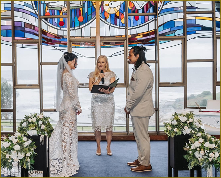 Bride and groom standing in front of an officiant during a wedding ceremony inside a chapel with stained glass windows and ocean view, decorated with floral arrangements.