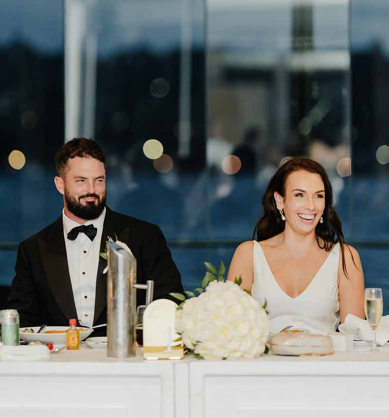 A bride and groom sitting at a reception table during their wedding celebration, with the bride smiling and the groom looking content. The table is decorated with a floral centerpiece and various drinks.