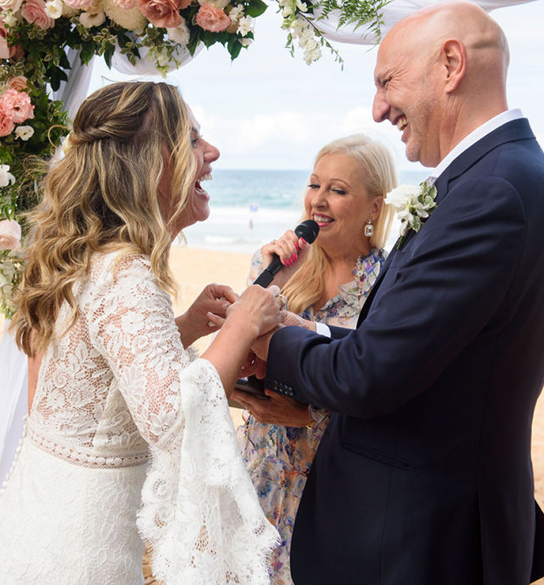 A beach wedding ceremony with a bride and groom exchanging vows under a floral arch, officiated by a woman holding a microphone.