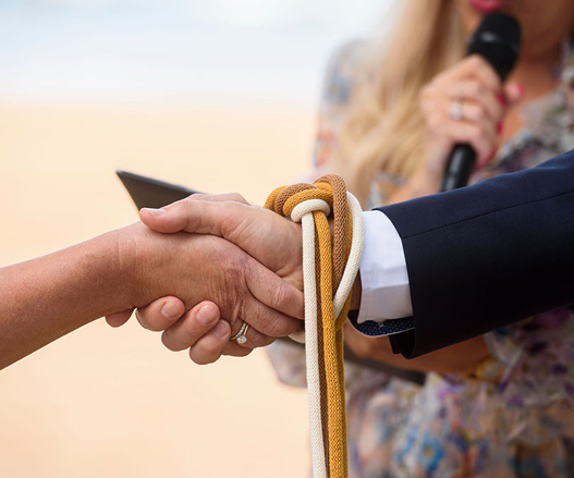 A handshake between two people, one in a suit, with a white shirt, and the other in a tan sleeve, with a rope tied around their hand. In the background, a woman is speaking into a microphone.