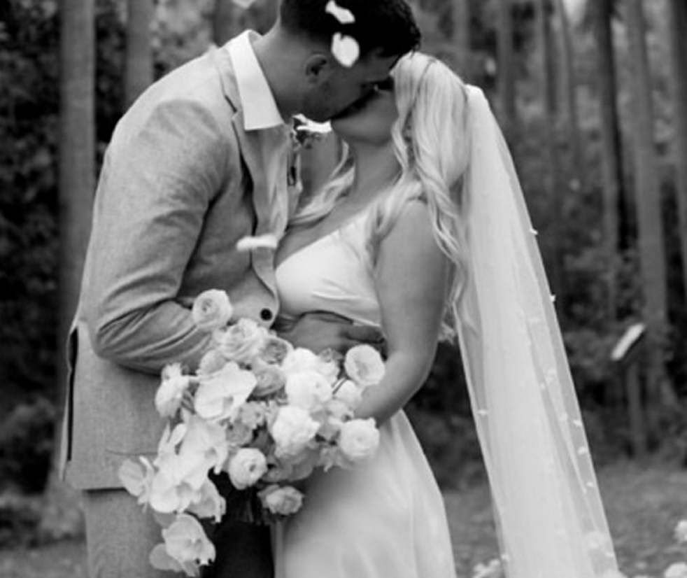 Black and white photo of a bride and groom sharing a kiss outdoors, with the groom holding a bouquet of flowers.