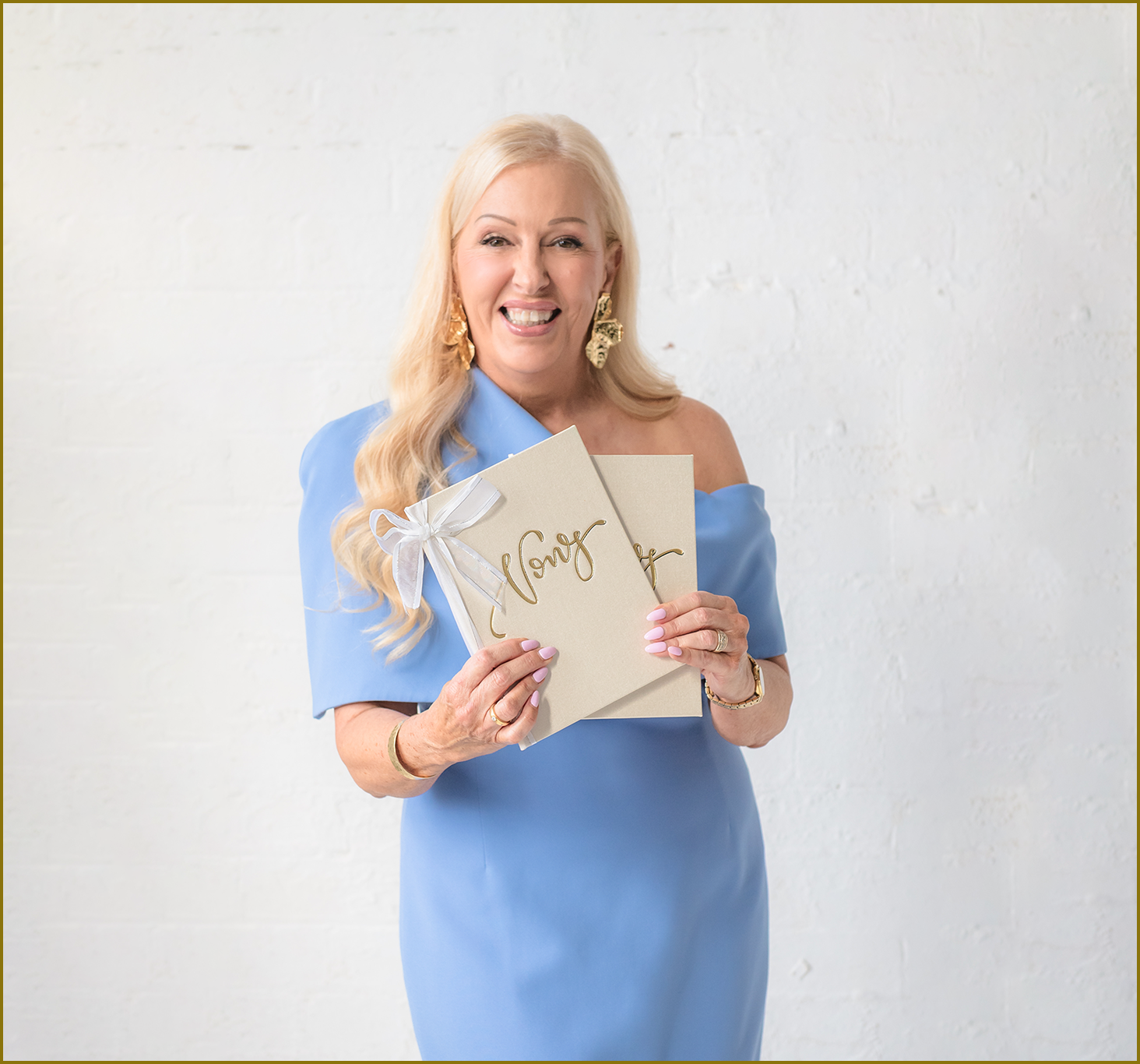 A woman with long blonde hair wearing a light blue off-the-shoulder dress and gold earrings holds a beige card with gold writing and a white ribbon against a white brick wall.