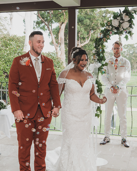 Bride and groom holding hands during wedding ceremony, with person in white suit standing nearby, decorated with flowers and greenery.