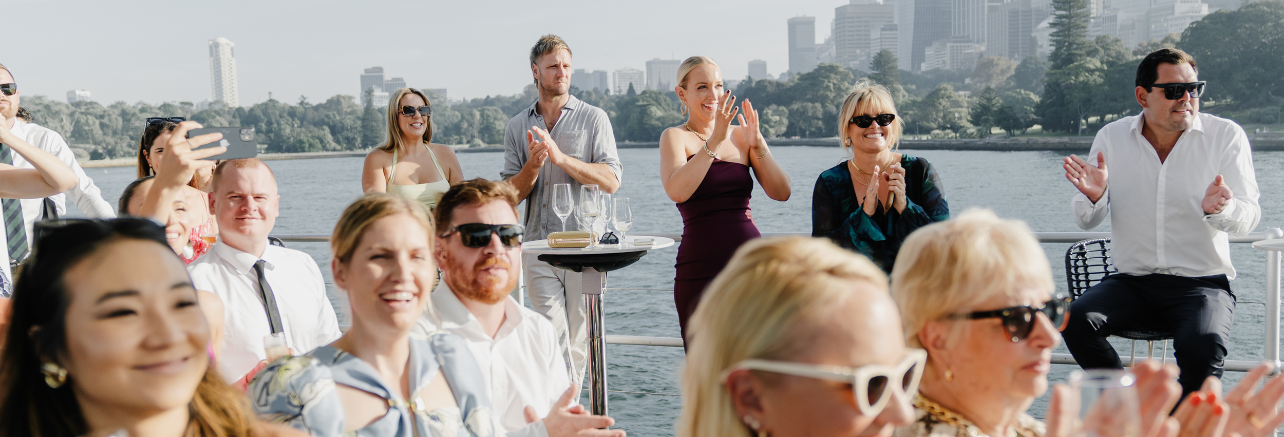 People enjoying a boat cruise by the water, some clapping and smiling, with a city skyline and trees in the background.
