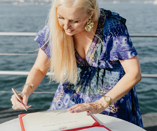 Woman with long blonde hair signing a document on a boat, water in the background.