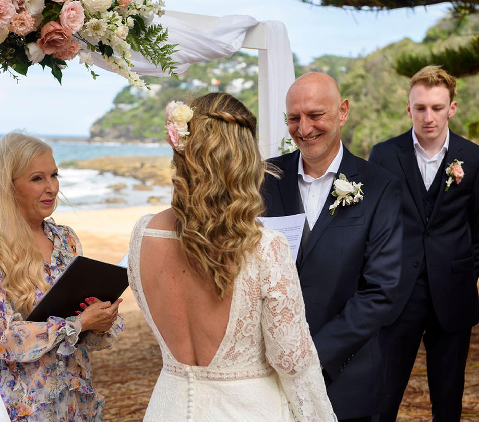 Bride and groom exchanging vows at an outdoor beach wedding with officiant and groomsmen.