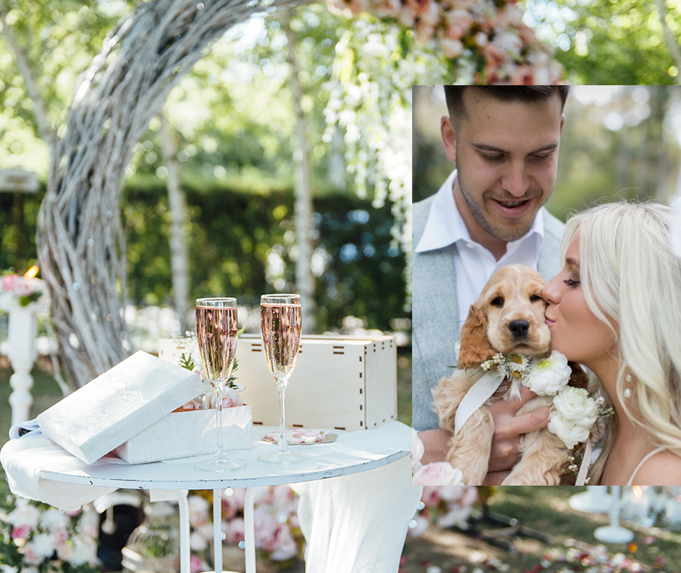 Champagne glasses, gift box, and flower arrangement on a small round table at an outdoor wedding reception with floral decor and a white arch in the background.
