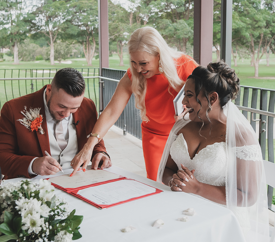 A bride and groom signing a marriage certificate at a table, with a woman in an orange dress assisting them during a wedding ceremony on a porch with a green park background.