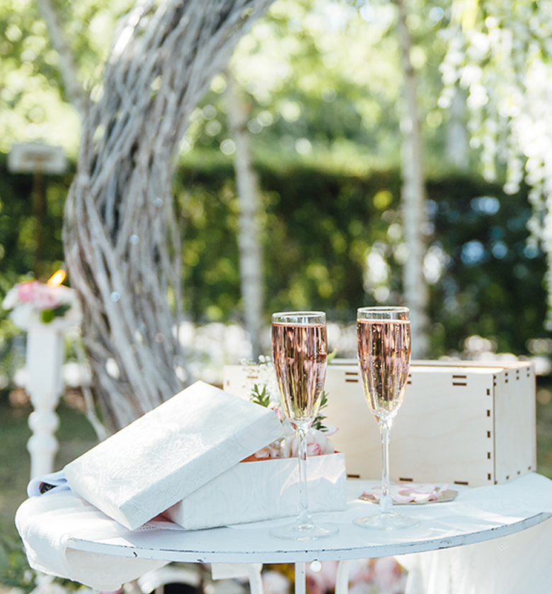 Two glasses of pink champagne on a white table outdoors, with wrapped gifts and a decorative arch in the background, suggesting a celebration or wedding.