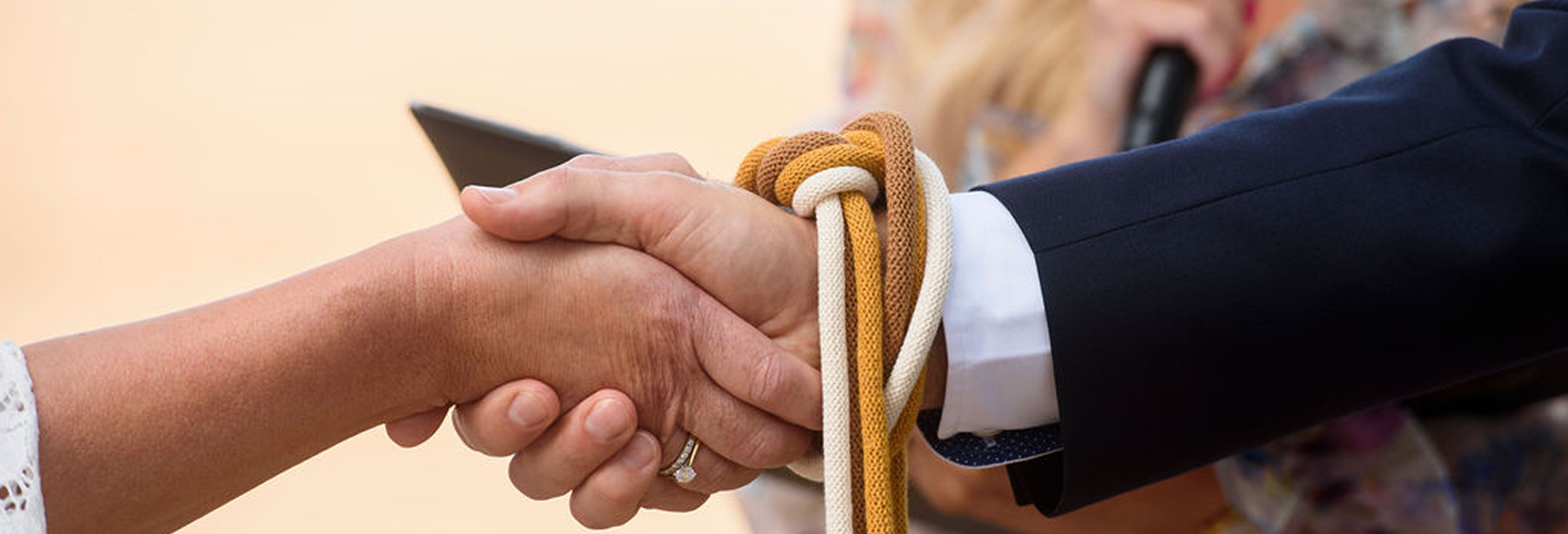 Close-up of a handshake between two women, one wearing a ring, with colorful ropes tied around the wrist of the person in a suit, during a formal ceremony.