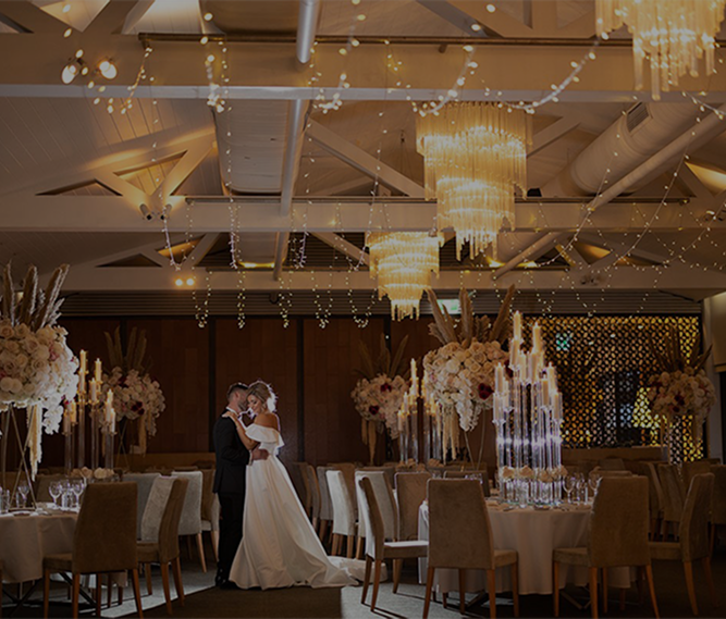 A bride and groom dancing in a decorated wedding reception hall with floral centerpieces and chandeliers.