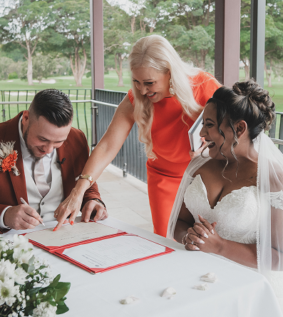 A couple signing a marriage certificate at their wedding, with a woman in an orange dress leaning over and smiling, and a woman in a white wedding dress sitting beside them.
