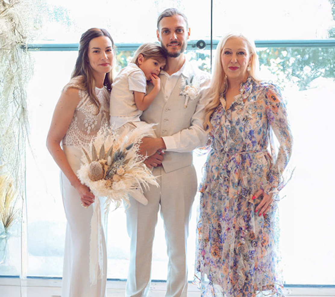 A family dressed in white and pastel colors celebrating at an indoor event with large windows showing a green outdoor scene in the background.