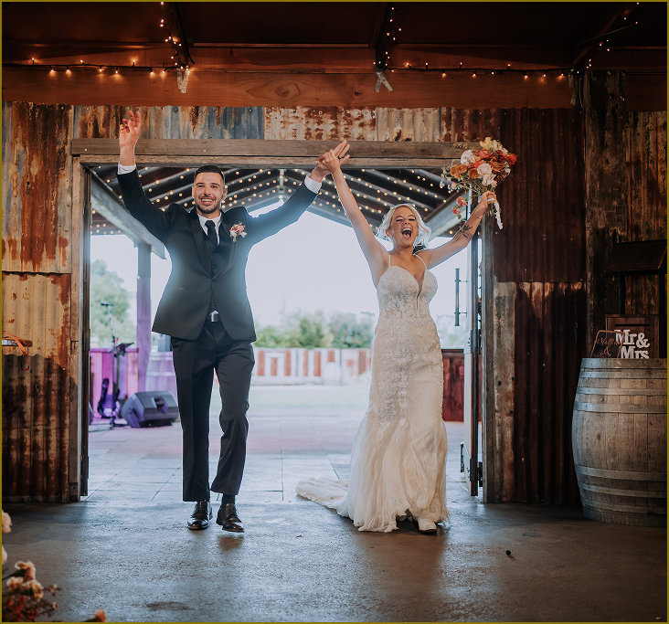 Wedding couple, bride in white gown and groom in black suit, celebrating with arms raised at reception venue.