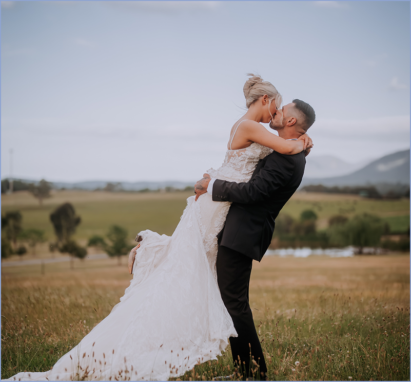 A couple in wedding attire embracing and kissing in a grassy field with mountains and trees in the background.