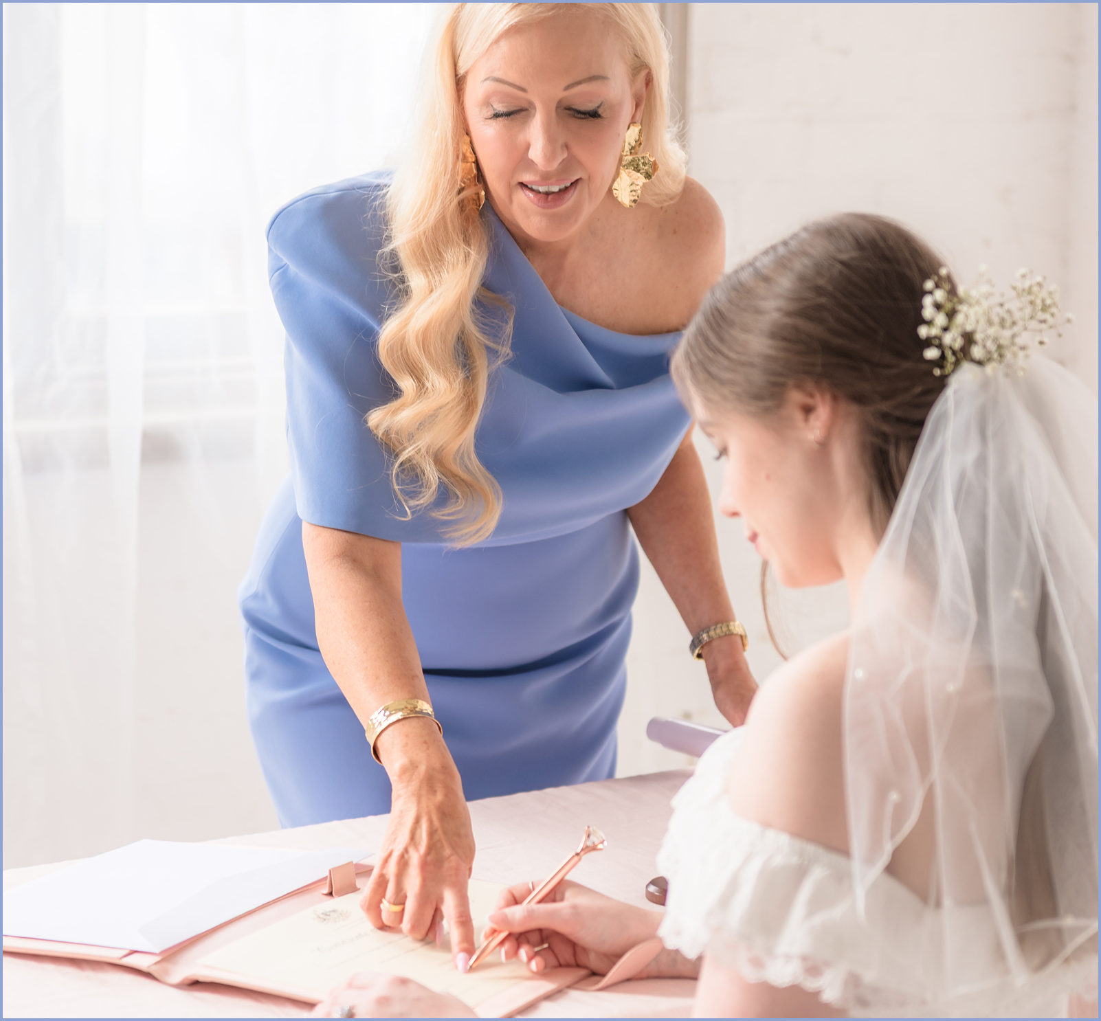 A woman with long blonde hair, dressed in a blue dress, is leaning over a table, looking at a young woman in a wedding dress who is sitting and signing a document. The young woman has a veil with white flowers and is holding a pink pen. The background is bright with white curtains.